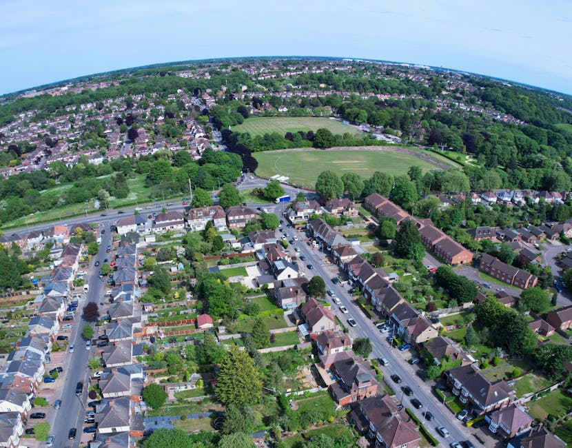 An aerial view of a suburban residential area featuring rows of terraced houses with small gardens, along with larger detached homes with lush lawns and neatly maintained hedges. The foreground shows 