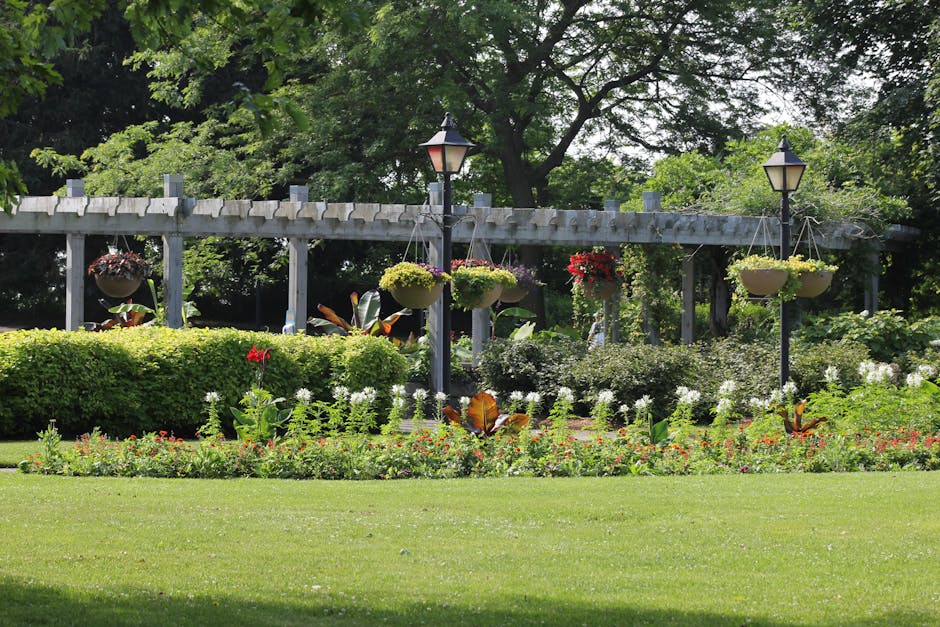 A well-maintained garden featuring a lush, neatly trimmed grass lawn in the foreground, bordered by colorful flowering plants and small shrubs. Behind the flower bed, there is a dense, rounded hedge w