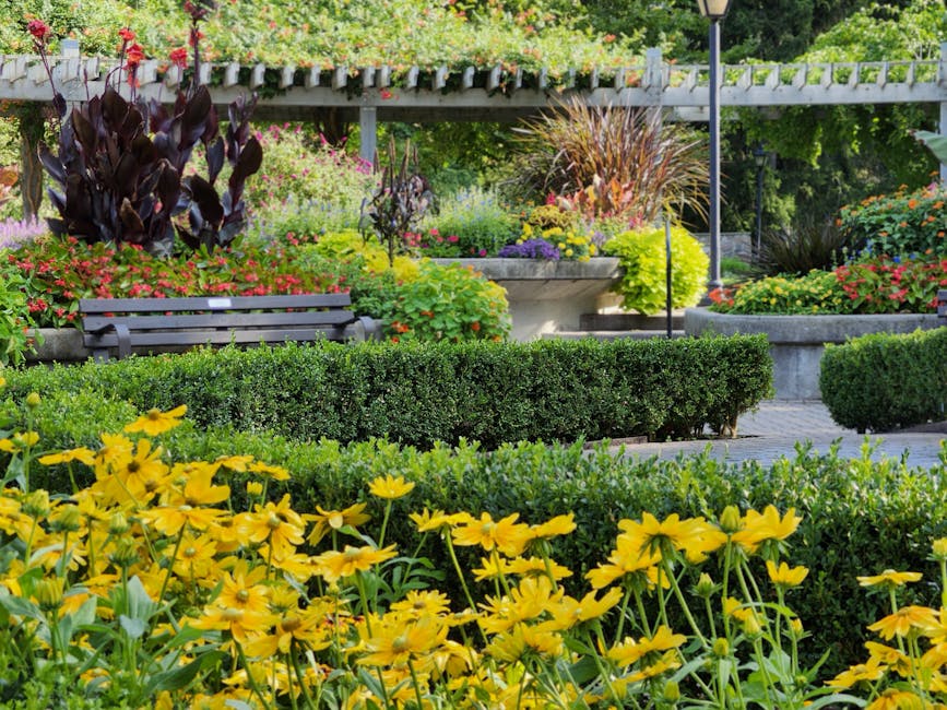 A well-maintained garden featuring a lush, neatly trimmed grass lawn in the foreground, bordered by colorful flowering plants and small shrubs. Behind the flower bed, there is a dense, rounded hedge w
